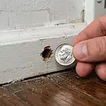 Quarter-inch mouse entry hole in wooden baseboard compared to the size of a US dime coin